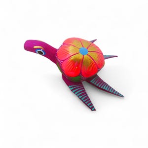 Może przedstawiać: Ręcznie wykonana, kolorowa figurka żółwia. Ciało żółwia jest w żywym różu, ze skorupą w kształcie kwiatu w kolorze czerwonym i złotym. Płetwy żółwia mają turkusowe paski. Głowa żółwia ma niebieskie oko.