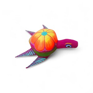 Może przedstawiać: Żywa, ręcznie malowana figurka żółwia. Skorupa żółwia ma jasny pomarańczowo-żółty kwiatowy wzór z turkusowym środkiem. Głowa i płetwy są w kolorze głębokiego różu, z turkusowymi i fioletowymi paskami na płetwach. Unikalny kawałek sztuki ludowej.