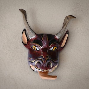 May include: A hand-painted mask featuring a dark red face with black accents, prominent curved horns, pointed ears, and wide yellow eyes. The mask displays a wide mouth with visible white teeth and a long, light-colored tongue protruding outward.
