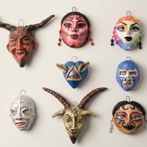 May include: A collection of eight decorative wall-hanging masks and ornaments featuring diverse designs, including horned figures, stylized faces with painted makeup, a blue heart with an eye, and metallic-colored wrestling-style masks.