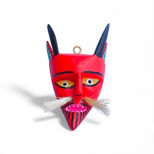 May include: A handcrafted, red devil mask with black horns and a white mustache. The mask features yellow eyes, a pink mouth, and black stripes on the chin. A gold loop is attached at the top for hanging. This decorative mask is perfect for Halloween or Day of the Dead.