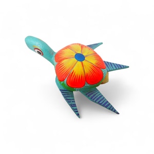 Może przedstawiać: Ręcznie malowana drewniana figurka żółwia. Żółw ma turkusowy korpus, ze skorupą zaprojektowaną jako kwiat w kolorze czerwonym, żółtym i pomarańczowym. Płetwy są turkusowe z niebieskimi paskami. Głowa żółwia ma jedno oko.