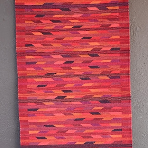 May include: A rectangular woven wall hanging featuring a vibrant gradient of red, orange, and purple horizontal stripes. The textile displays a repeating geometric pattern of slanted dashes throughout, finished with white fringe at the top and bottom edges.