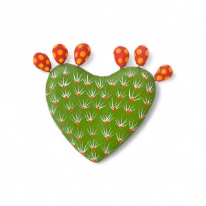 Puede incluir: Un cactus verde en forma de corazón con detalles blancos y rojos, y seis frutos rojos y naranjas. El cactus está pintado con un acabado brillante y se encuentra sobre un fondo blanco. El diseño es una representación caprichosa de un nopal.