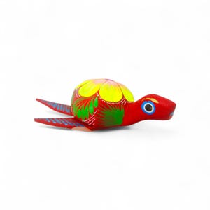 Może przedstawiać: Ręcznie malowana drewniana figurka żółwia. Żółw ma czerwone ciało i głowę, z żółtą i różową skorupą ozdobioną zielonymi i białymi wzorami kwiatowymi. Oczy są niebieskie z białym konturem. Płetwy żółwia są czerwone z niebieskimi akcentami.