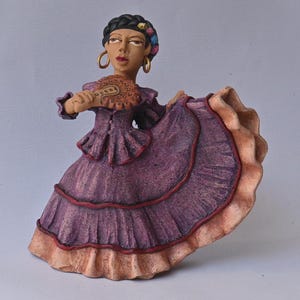 Puede incluir: Una figura de arcilla de una persona con cabello oscuro trenzado con detalles coloridos, que lleva pendientes de aro dorados y un vestido morado con falda de volantes y dobladillo color melocotón. Sostiene un abanico redondo y texturizado.