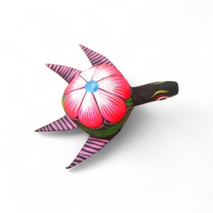 Może przedstawiać: Ręcznie malowana drewniana figurka żółwia z kwiatowym wzorem. Skorupa to duży różowy kwiat z niebieskim środkiem. Płetwy są w paski w kolorze różowym i czarnym. Głowa jest czarna z żółtymi i czerwonymi akcentami.