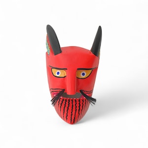 May include: A red devil mask with black horns, yellow eyes with blue pupils, and a black mustache and beard. The beard has vertical black stripes. The mask is likely made of wood and is a decorative item.