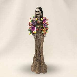 May include: A standing skeletal figure with a white skull face and dark hair, wearing a textured brown gown that resembles tree bark. The figure is adorned with a vibrant floral arrangement featuring purple, yellow, and red flowers around the shoulders.