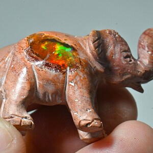 May include: A small carved elephant figurine made of red jasper with a natural opal embedded in its back. The opal displays a rainbow of colors, including red, orange, yellow, green, and blue.