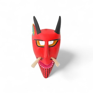 May include: A red devil mask with black horns, yellow-lined eyes, and a long nose. The mask has a wide, open mouth with pink and white teeth, and two straw-like whiskers. The mask is likely used for costume or decorative purposes.