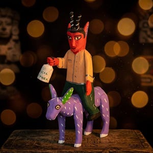 May include: A hand-carved wooden figurine of a red-skinned devil with black horns, wearing a beige shirt and green pants, riding a purple donkey with white markings. The devil holds a white bottle labeled "CAL CHUGA" in its right hand.