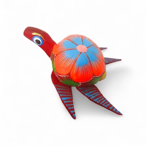 Może przedstawiać: Ręcznie malowana drewniana figurka żółwia. Żółw ma czerwone ciało i głowę, z niebiesko-pomarańczowym pancerzem przypominającym kwiat. Nogi i głowa mają niebieskie paski. Oczy są niebieskie z czarnymi źrenicami.