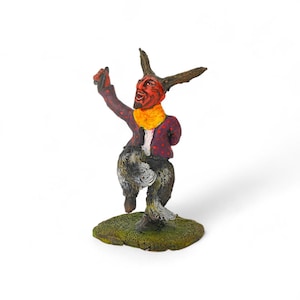 May include: A whimsical figurine of a devil-like creature with a red face, horns, and a yellow scarf. The figure wears a purple jacket with red polka dots, gray fur-like pants, and stands on a green, grassy base. The figure is posed with one arm raised.