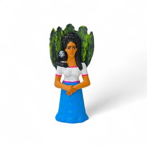 May include: A hand-painted ceramic figurine of a woman with long black hair, wearing a white top with pink trim and a blue skirt. A small monkey sits on her shoulder, and a green leaf-shaped element is behind her.