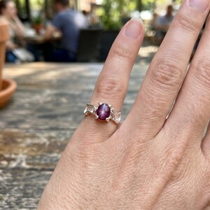 May include: A silver ring featuring a central oval-shaped deep pink star gemstone. The band has a decorative cutout design on both sides of the stone setting. The gemstone displays a distinct four-rayed star pattern across its surface.