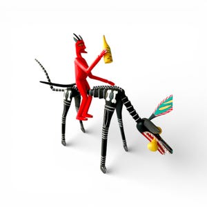 May include: A whimsical, hand-painted wooden sculpture featuring a red devil figure riding a black and white dog-like creature. The devil holds a yellow bottle. The dog has a detailed design with bone accents and colorful accents on the ears and mouth.