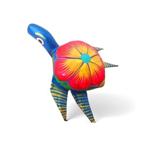 Może przedstawiać: Żywa, ręcznie malowana drewniana figurka żółwia. Żółw ma niebieską głowę i nogi z żółtymi paskami oraz skorupę ozdobioną dużym czerwono-żółtym kwiatowym wzorem. Unikalny element sztuki ludowej.