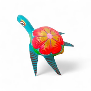 Może przedstawiać: Ręcznie malowana drewniana figurka żółwia. Żółw pomalowany jest na jasny turkus, czerwony, złoty i zielony. Skorupa ma duży czerwony kwiat ze złotymi akcentami. Nogi i głowa są turkusowe z brązowymi paskami. Unikalny kawałek sztuki ludowej.