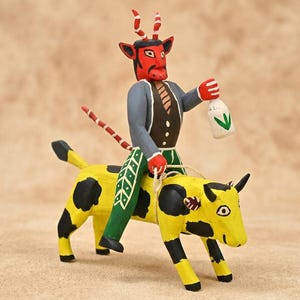 May include: A hand-painted wooden figurine of a red-faced devil with striped horns and a tail, wearing a grey jacket and green pants, riding a yellow cow with black spots. The devil holds a white bottle with a green leaf design and text.