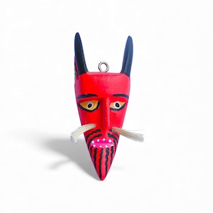 May include: A red, hand-painted wooden mask with black horns and a white mustache. The mask features yellow eyes, a pink mouth, and black stripes on the chin. A small metal loop is at the top for hanging.