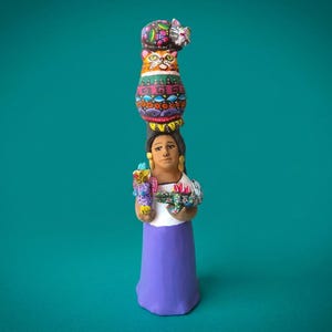 Puede incluir: Una figura de arcilla pintada a mano de una persona con blusa blanca y falda morada, sosteniendo un pájaro y un lagarto coloridos. Sobre la cabeza de la persona hay una pila de dos gatos con intrincados patrones geométricos y florales.