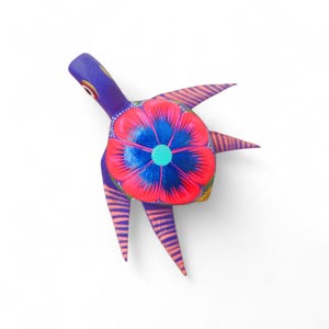 Może przedstawiać: Żywo pomalowana drewniana figurka żółwia. Żółw ma fioletową głowę i nogi, czerwono-niebieski wzór kwiatowy na grzbiecie oraz pomarańczowo-różowe paski. Dzieło sztuki ludowej jest umieszczone na białym tle.