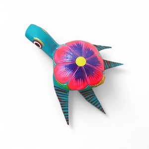 Może przedstawiać: Ręcznie malowana drewniana figurka żółwia. Żółw jest pomalowany na turkusowo z dużym kwiatowym wzorem na grzbiecie, z różowymi i fioletowymi płatkami i żółtym środkiem. Nogi żółwia mają brązowe paski.