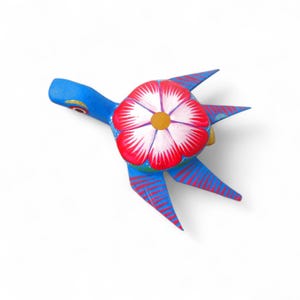 Może przedstawiać: Ręcznie malowana drewniana figurka żółwia. Żółw jest głównie niebieski z czerwonymi i białymi akcentami. Skorupa to duży kwiat z czerwonymi płatkami i białymi detalami. Płetwy żółwia mają czerwone paski.