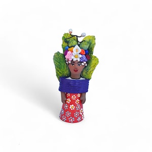 May include: A hand-painted ceramic figurine of a woman with dark skin, wearing a red dress with white and blue floral patterns. She has a blue shawl and a floral headdress with green leaves. The figurine is approximately 5 inches tall.