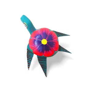 Może przedstawiać: Ręcznie malowana drewniana figurka ryby. Ryba jest turkusowa z czerwonymi i niebieskimi paskami oraz dużym kwiatem z czerwonymi, fioletowymi i żółtymi akcentami. Rękodzieło na białym tle.