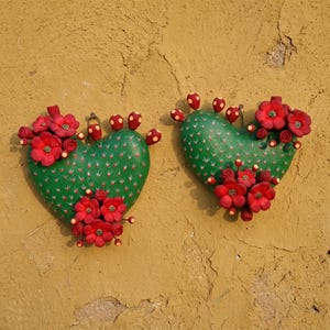 Puede incluir: Dos decoraciones de pared con forma de corazón que representan cactus verdes con flores rojas y pequeños frutos rojos con puntos amarillos. Cada pieza tiene una superficie texturizada con pequeñas espinas y racimos de flores rojas.