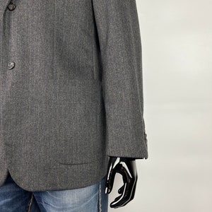 May include: A dark gray blazer with a subtle herringbone pattern. The blazer has a two-button closure and a flap pocket on the left side.