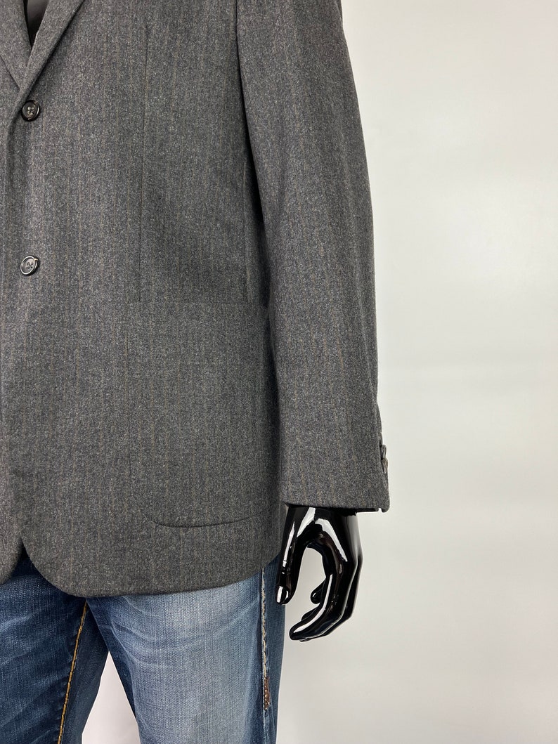 May include: A dark gray blazer with a subtle herringbone pattern. The blazer has a two-button closure and a flap pocket on the left side.