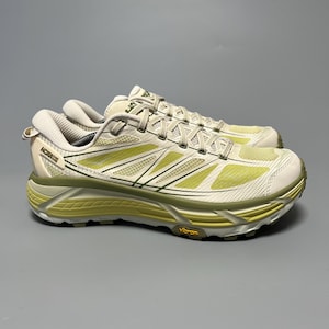Könnte beinhalten: Paar Hoka-Laufschuhe in Creme, Hellgrün und Olivgrün. Die Schuhe haben eine dicke Sohle mit Vibram-Branding und ein atmungsaktives Mesh-Obermaterial. Die Schnürsenkel und das Hoka-Logo sind auf der Seite des Schuhs sichtbar. Der Modellname ist sichtbar.