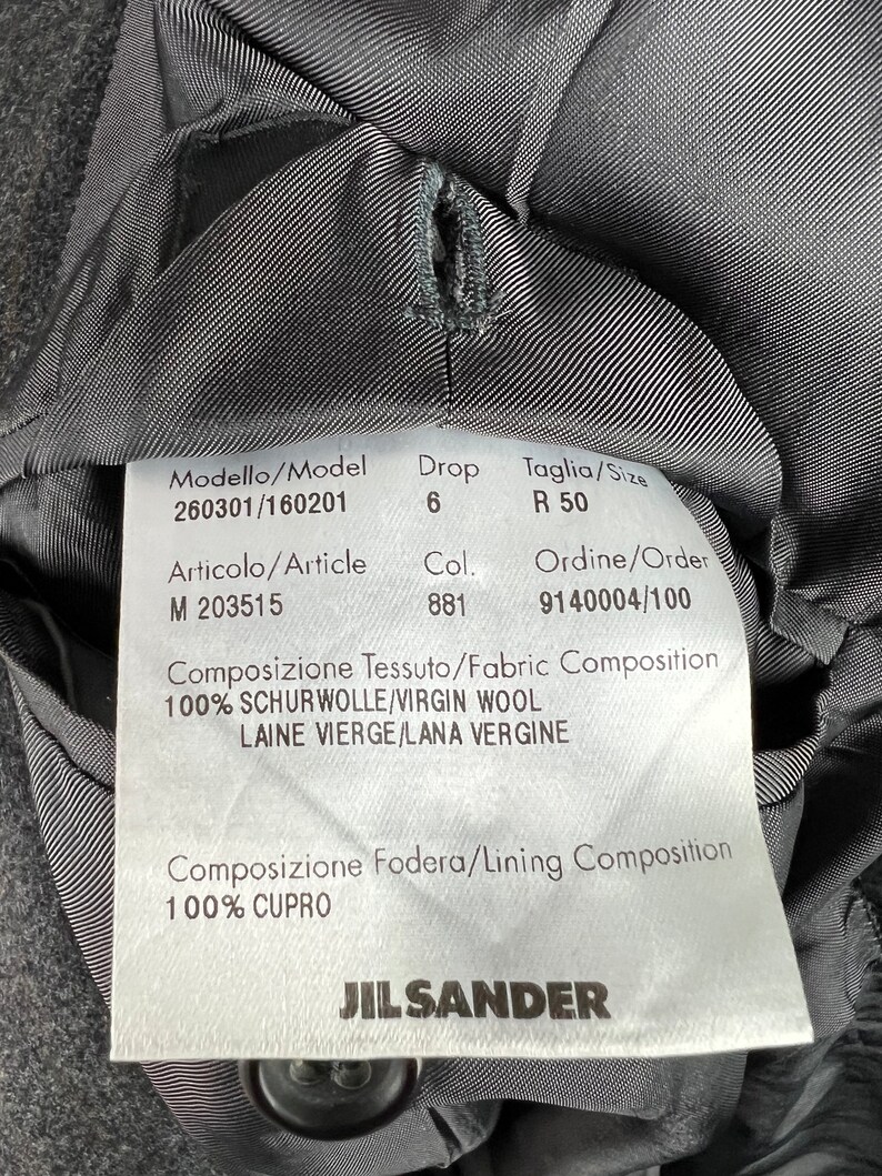 May include: A black Jil Sander clothing label with white text. The label includes the model number 260301/160201, drop size 6, size R 50, article number M 203515, color 881, order number 9140004/100, fabric composition 100% virgin wool, and lining composition 100% cupro.