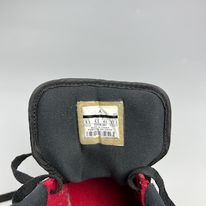 Puede incluir: Primer plano del interior de una zapatilla deportiva negra y roja. La leng&uuml;eta del zapato presenta una etiqueta con informaci&oacute;n sobre la talla y la fabricaci&oacute;n, incluyendo la talla US 9.5, la talla EUR 43 y la fecha de fabricaci&oacute;n 01/03/14.