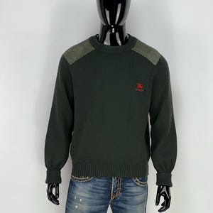 May include: Dark green sweater with suede shoulder patches and ribbed cuffs. Features a small red logo on the chest. The sweater is displayed on a mannequin, paired with blue jeans.