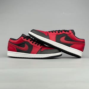 Puede incluir: Un par de zapatillas bajas rojas y negras con suela blanca. Las zapatillas tienen un logotipo de swoosh negro en el lateral y cordones negros. Los zapatos se muestran sobre un fondo gris.