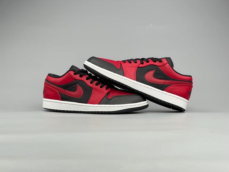 Puede incluir: Un par de zapatillas bajas rojas y negras con suela blanca. Las zapatillas tienen un logotipo de swoosh negro en el lateral y cordones negros. Los zapatos se muestran sobre un fondo gris.