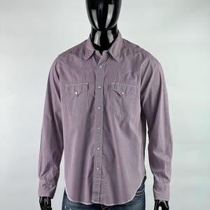 Puede incluir: Una camisa de manga larga con botones y un patrón a cuadros en tonos de rosa y blanco. La camisa tiene un cuello puntiagudo, dos bolsillos en el pecho y un puño de un solo botón.