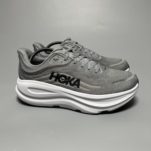 May include: A pair of grey athletic sneakers featuring a breathable mesh upper, a thick white cushioned midsole, and the black HOKA logo printed on the side. The shoes include grey laces and a padded collar, designed with a low-top silhouette.