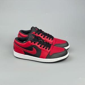 Puede incluir: Un par de zapatillas bajas rojas y negras con suela blanca. Las zapatillas presentan una parte superior roja con detalles en negro, incluyendo los cordones, el cuello y un logotipo de swoosh en el lateral. Las zapatillas est&aacute;n sobre un fondo gris.