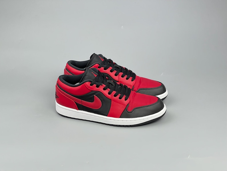 Puede incluir: Un par de zapatillas bajas rojas y negras con suela blanca. Las zapatillas presentan una parte superior roja con detalles en negro, incluyendo los cordones, el cuello y un logotipo de swoosh en el lateral. Las zapatillas est&aacute;n sobre un fondo gris.