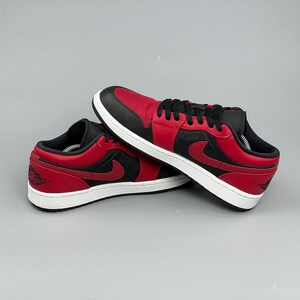 Puede incluir: Un par de zapatillas bajas rojas y negras con suelas blancas. Las zapatillas tienen una puntera, cordones y cuello negros, con un cuerpo rojo y un logotipo de swoosh negro en el lateral. Los zapatos se muestran sobre un fondo gris.