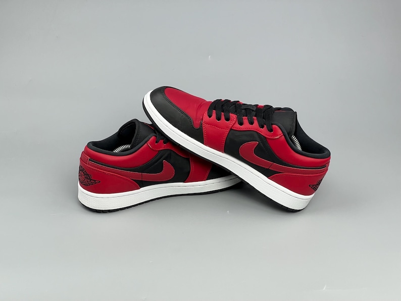 Puede incluir: Un par de zapatillas bajas rojas y negras con suelas blancas. Las zapatillas tienen una puntera, cordones y cuello negros, con un cuerpo rojo y un logotipo de swoosh negro en el lateral. Los zapatos se muestran sobre un fondo gris.