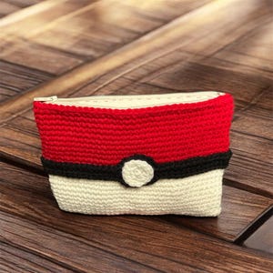 Puede incluir: Una bolsa de cremallera de crochet roja, blanca y negra con un diseño de Poké Ball.
