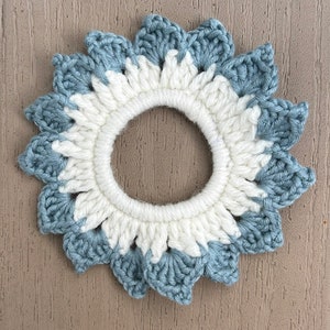 May include: A crocheted hair scrunchie in a light blue and white color scheme. The scrunchie is shaped like a flower with a white center and blue petals.
