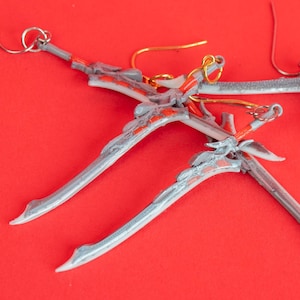 Può includere: Orecchini unici fatti a mano con un design a forma di spada. Gli orecchini presentano una base grigio-argento con dettagli rossi e arancioni. I ganci per le orecchie sono color oro. Su sfondo rosso brillante.