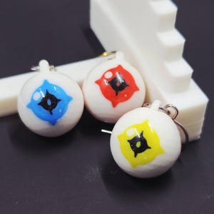 May include: Three white, spherical charms with eye designs in blue, red, and yellow. Each charm has a black pupil and a silver-colored keyring. The charms are displayed near a white, stepped structure against a dark background.